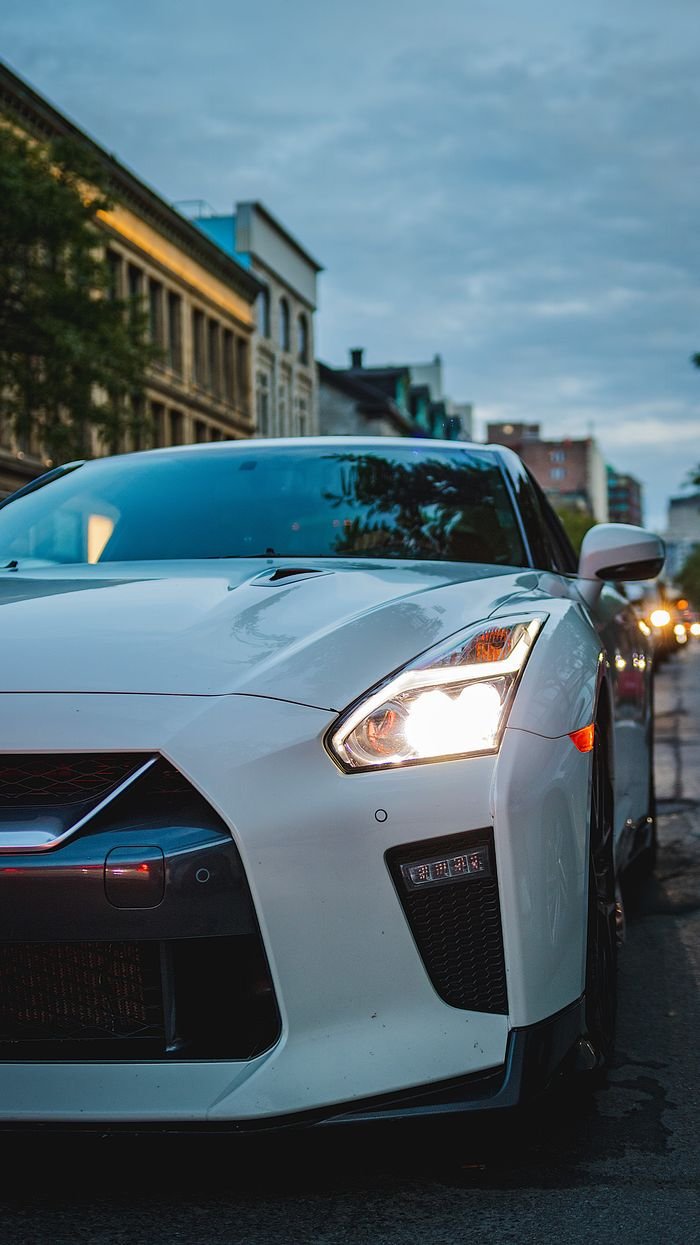 White sports car with headlights on, parked on a city street at dusk.